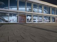 an empty plaza with large windows and people walking along it outside of it at night