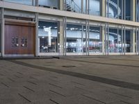 an empty plaza with large windows and people walking along it outside of it at night