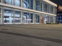 an empty plaza with large windows and people walking along it outside of it at night