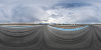 an empty racetrack that is grey with blue trims and clouds overhead it is seen in a fish eye lens