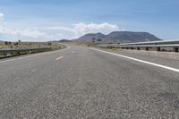 an empty roadway with hills and mountains in the background under a blue sky with clouds