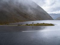 an empty paved road leads down to a lake with mountains in the background in the clouds