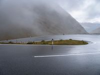 an empty paved road leads down to a lake with mountains in the background in the clouds