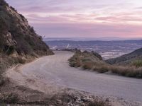 an empty road leading to the mountain above a city at sunset by the ocean with mountains and palm trees