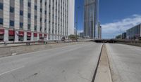 an empty road between two high rise buildings with the top of a freeway crossing to its side