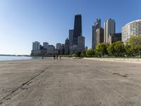 the empty road is beside water and tall buildings in the background with people walking around it