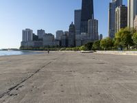 the empty road is beside water and tall buildings in the background with people walking around it