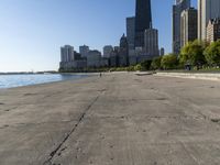 the empty road is beside water and tall buildings in the background with people walking around it