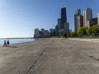 the empty road is beside water and tall buildings in the background with people walking around it