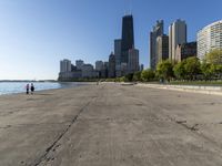 the empty road is beside water and tall buildings in the background with people walking around it