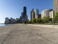 the empty road is beside water and tall buildings in the background with people walking around it