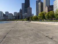 the empty road is beside water and tall buildings in the background with people walking around it