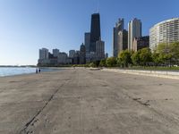 the empty road is beside water and tall buildings in the background with people walking around it