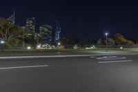 an empty road with city lights behind it at night, in the background is a row of green buildings