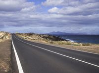 an empty road on a deserted beach with mountains in the background and a cloudy sky