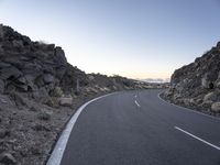 an empty winding road on a mountain with the sun set above the mountains in the background