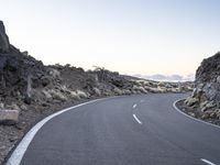 an empty winding road on a mountain with the sun set above the mountains in the background