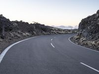 an empty winding road on a mountain with the sun set above the mountains in the background