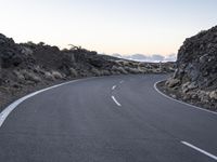 an empty winding road on a mountain with the sun set above the mountains in the background