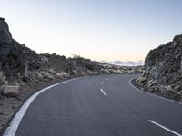 an empty winding road on a mountain with the sun set above the mountains in the background