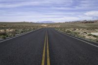 empty road in the middle of an open desert area with hills in the distance, and yellow lines of the asphalt on one side