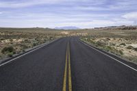 empty road in the middle of an open desert area with hills in the distance, and yellow lines of the asphalt on one side