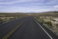 empty road in the middle of an open desert area with hills in the distance, and yellow lines of the asphalt on one side