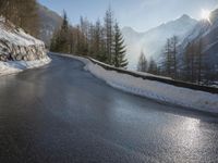 an empty road near some mountains and snow covered ground with the sun behind it with trees and hills in front