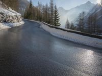 an empty road near some mountains and snow covered ground with the sun behind it with trees and hills in front