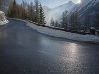 an empty road near some mountains and snow covered ground with the sun behind it with trees and hills in front