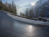 an empty road near some mountains and snow covered ground with the sun behind it with trees and hills in front