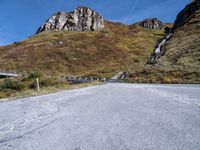 an empty road has rocks and gravel beside it on a hillside of grass and rock with mountains