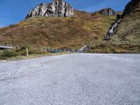 an empty road has rocks and gravel beside it on a hillside of grass and rock with mountains