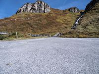 an empty road has rocks and gravel beside it on a hillside of grass and rock with mountains