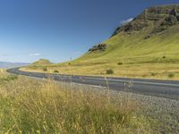 an empty road passing through the mountains with yellow flowers in a grassy field, on a clear day