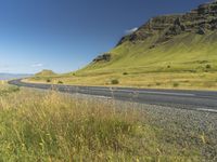 an empty road passing through the mountains with yellow flowers in a grassy field, on a clear day