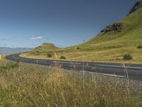 an empty road passing through the mountains with yellow flowers in a grassy field, on a clear day