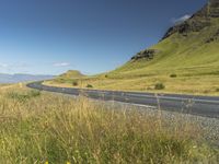 an empty road passing through the mountains with yellow flowers in a grassy field, on a clear day