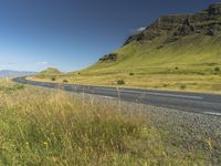 an empty road passing through the mountains with yellow flowers in a grassy field, on a clear day