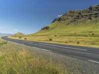 an empty road passing through the mountains with yellow flowers in a grassy field, on a clear day