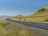 an empty road passing through the mountains with yellow flowers in a grassy field, on a clear day