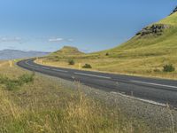 an empty road passing through the mountains with yellow flowers in a grassy field, on a clear day