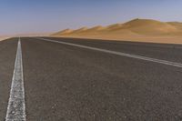 a wide empty road with two lines on the pavement, in front of sand dunes