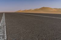 a wide empty road with two lines on the pavement, in front of sand dunes