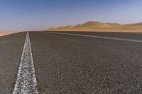 a wide empty road with two lines on the pavement, in front of sand dunes