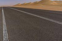 a wide empty road with two lines on the pavement, in front of sand dunes