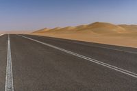 a wide empty road with two lines on the pavement, in front of sand dunes