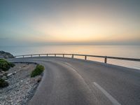 an empty road curves into the distance of some mountains with ocean in background and sun setting