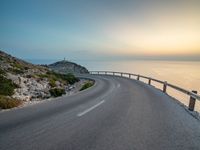 an empty road curves into the distance of some mountains with ocean in background and sun setting