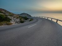 an empty road curves into the distance of some mountains with ocean in background and sun setting